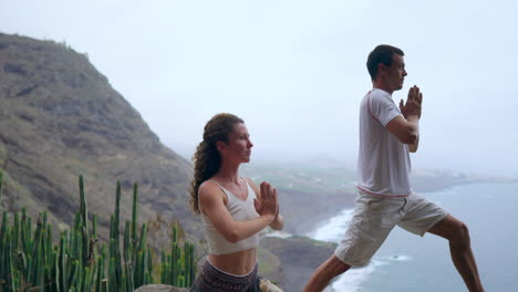 Ein-Mann-Und-Eine-Frau,-Die-Am-Rand-Einer-Klippe-Mit-Blick-Auf-Das-Meer-Stehen,-Heben-Ihre-Hände-Und-Atmen-Beim-Yoga-Die-Meeresluft-Ein
